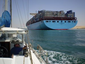 A small sailboat follows a huge freighter with a beam of about fifty feet